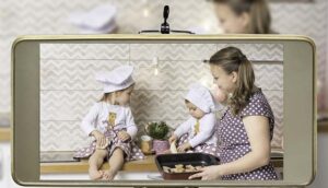 Young female blogger and vlogger online influencer mom with two daughters, live streaming a cooking, baking cookies with children show on social media using a smartphone.