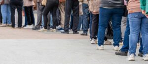 En grupp människor står i kö utomhus på en asfalterad eller stenlagd yta. Bilden är tagen i benhöjd och visar främst personernas underkroppar och skor. Kön sträcker sig längs bilden från höger till vänster, och personerna bär vardagskläder som jeans, byxor och jackor i olika färger. Ingen ansiktet syns.